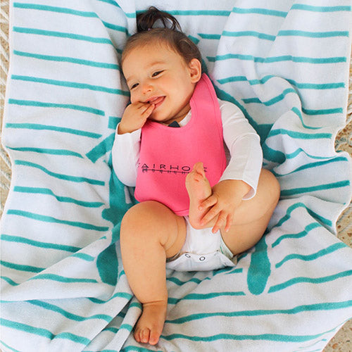 Smiling baby lying on a beach towel while wearing a pink bib with the word 